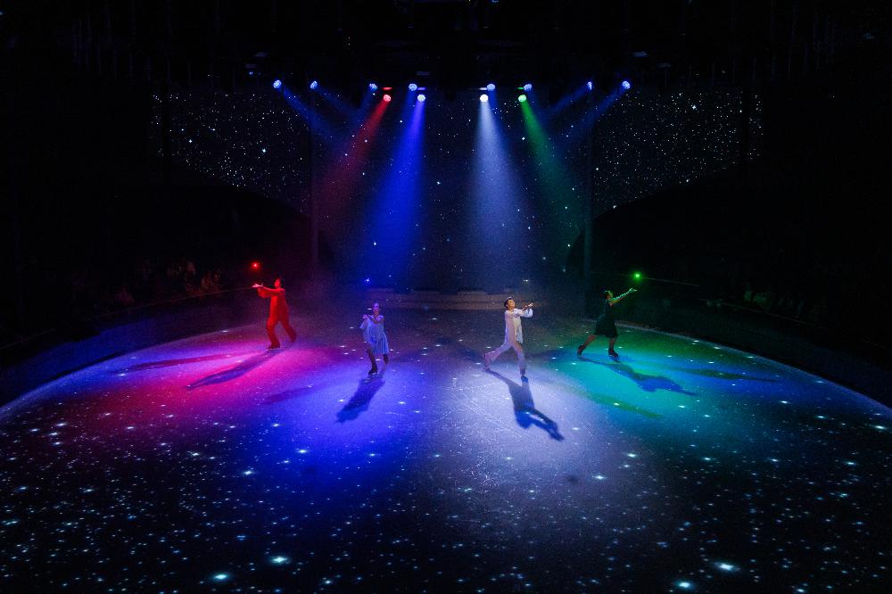 Four ice skaters performing under coloured spotlights on a starlit rink